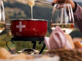 A red fondue caquelon sits on a table in the outdoor seating area. Melted cheese is scooped out with forks. Wine glasses and a bread basket are placed next to it. Wooded slopes and Alpine mountains are visible in the background.