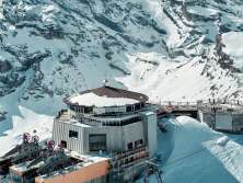 The drone shot shows the Schilthorn with the distinctive Piz Gloria revolving restaurant and the mountain station, nestled in the snow-covered high-mountain landscape. The snow-capped mountain ranges of the Bernese Alps stretch out around the summit, lending the scene its impressive vastness.