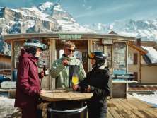 Three people raise their glasses together at the Gondelbar near the Hotel Jungfrau in Mürren. The cozy terrace, with its view of the snow-covered mountain landscape, is the perfect place to relax after a day on the slopes.