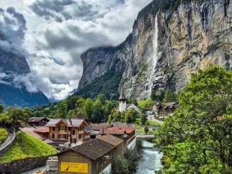 A view of the village of Lauterbrunnen with its chalets and the flowing Lütschine River. In the background, the Staubbach Falls plunge from the steep rock face into the valley, defining the village’s skyline.