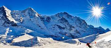 A groomed ski slope winds through the snow-covered high-mountain landscape in the foreground. Behind it, the striking panorama of the Eiger, Mönch, and Jungfrau stretches out under a clear sky, illuminated by the low-hanging sun.