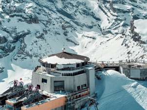 The drone shot shows the Schilthorn with the distinctive Piz Gloria revolving restaurant and the mountain station, nestled in the snow-covered high-mountain landscape. The snow-capped mountain ranges of the Bernese Alps stretch out around the summit, lending the scene its impressive vastness.