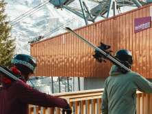 Three people carry their skis to the Mürren station of the Schilthornbahn. Passing the modern station buildings and with a view of the snow-covered mountain landscape, they begin their day of skiing.