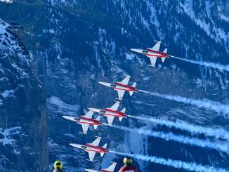 Planes from the Patrouille Suisse fly in tight formation over the Lauberhorn Race. In the foreground, spectators stand on a snow-covered slope and watch the air show as the planes leave trails of smoke against the steep, snow-covered mountainside.