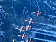 Planes from the Patrouille Suisse fly in tight formation over the Lauberhorn Race. In the foreground, spectators stand on a snow-covered slope and watch the air show as the planes leave trails of smoke against the steep, snow-covered mountainside.