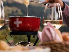 A red fondue caquelon sits on a table in the outdoor seating area. Melted cheese is scooped out with forks. Wine glasses and a bread basket are placed next to it. Wooded slopes and Alpine mountains are visible in the background.
