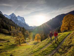 Two hikers are out and about in the Rosenlaui Valley countryside. There’s an autumnal atmosphere, and the sky is covered with light, hazy clouds. The mountain panorama is blanketed in snow.