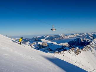 A skier carves turns on the slope at the Mürren-Schilthorn ski resort. The Schilthornbahn gondola takes skiers to the summit. A snow-covered mountain panorama can be seen in the background.