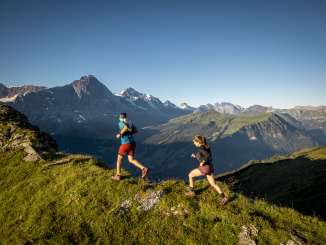 Two trail runners are running along a mountain ridge with a view of the Männlich and the Eiger North Face.