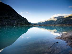 Lake Gelmersee is nestled in the high-alpine landscape of the Grimsel region. The calm surface of the water reflects the surrounding mountain slopes and the clear sky. The rocky shores are visible in the foreground, while the dam borders the lake in the background.