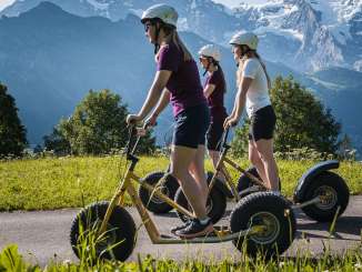 With the snow-capped mountains in the background, three women on scooters ride along a paved path surrounded by meadows and trees.