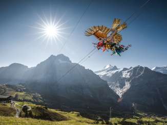 The First Glider flies over a green meadow, with mountains rising in the background beneath a clear blue sky.
