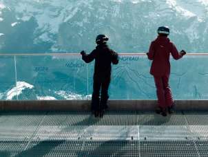 Two women in ski gear are standing at the railing of the Skyline Walk platform, enjoying the view of the Eiger, Mönch, and Jungfrau.