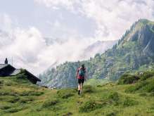 Eine Wanderin steigt über eine grüne Alpweide oberhalb der Lobhornhütte. Links ist ein Teil der Hütte sichtbar, rechts erhebt sich ein steiler, bewachsener Felsrücken. Wolken ziehen durch die umliegende Berglandschaft.