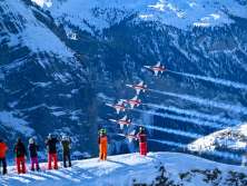 Planes from the Patrouille Suisse fly in tight formation over the Lauberhorn Race. In the foreground, spectators stand on a snow-covered slope watching the air show, while the planes leave trails of smoke against the steep, snow-covered mountainside.