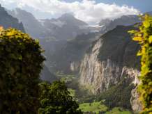 Der Blick führt über steile Felswände und grüne Talhänge tief ins Lauterbrunnental. Im Hintergrund erheben sich schroffe Berggipfel wie das Breithorn und Gletscher, teilweise von Wolken umgeben.