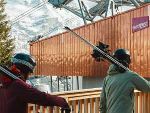 Three people carry their skis to the Mürren station of the Schilthornbahn. Passing the modern station buildings and with a view of the snow-covered mountain landscape, they begin their day of skiing.