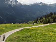 Two cyclists follow the winding Grosse Scheidegg Pass road through lush green alpine meadows. The view stretches across the Grindelwald Valley to the Mättenberg and the Eiger, while thick clouds create a striking atmosphere.